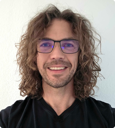 A man with a light beard wearing black glasses with a purple tint and a black v-necked t-shirt smiles in front of a white wall.