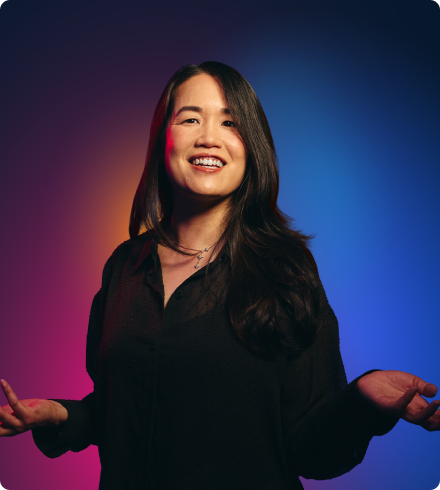 An Asian woman wearing a black blouse and a necklace smiles brightly with her arms spread out to the side and her hands held up.