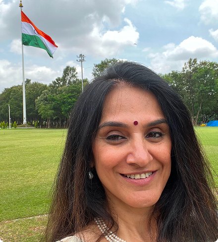 An Indian woman with long dark hair, a bindhi, and a multi-strand of pearls on  stands outside in front of a lawn with an Indian flag and trees in the background.