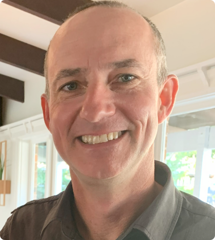 A white man with a receding hairline wears a brown collared shirt and smiles directly at the camera in front a bank of windows in a house.