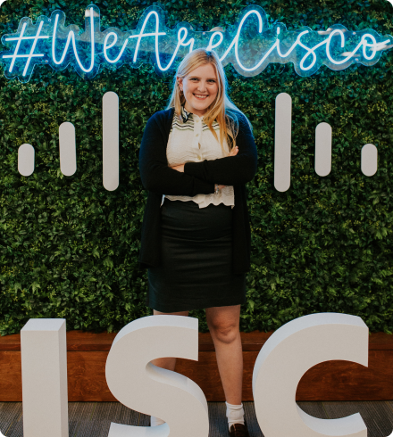 A woman wearing a black skirt and white blouse with black and green angled stripes at the top under a black sweater stands with her arms crossed in front of a leaf-covered wall with a neon sign that says "We Are Cisco."