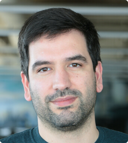 A dark-haired man with a scruffy beard wearing a t-shirt looks seriously at the camera in front of a blurred background.