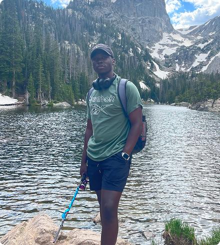 Tanaka holds a fishing rod and wears a backpack standing in front of a picturesque mountain lake.