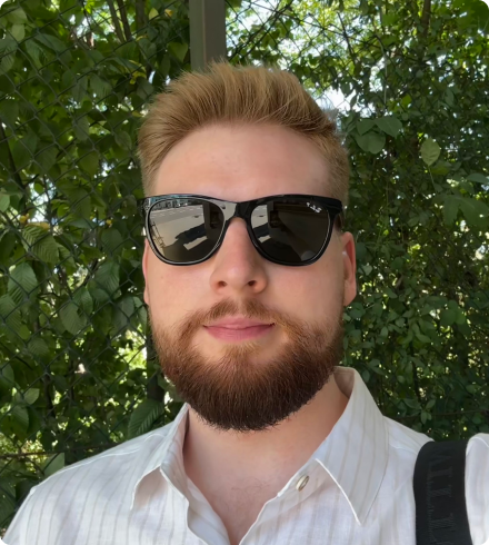 Tomasz looks at the camera while wearing sunglasses in front of a leafy background.