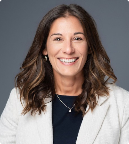 A woman with dark shoulder-length hair wearing a blue blouse under a white suit jacket with a silver necklace smiles directly at the camera in front of a gray background.