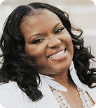 A Black woman wearing white feather earrings, a white blazer, and a white two-strand stone necklace tilts her head and smiles.