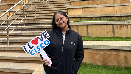 A woman with glasses stands in front of a staircase holding a sign that says, " I Love Cisco."
