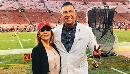 A woman wearing a baseball cap and a man in a suit jacket holding up two fingers in a V stand on a field in a stadium.