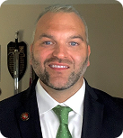 A man with graying hair and a beard wears a white button-down shirt with a green tie and black suit jacket while standing in front of a beige wall with a sword and shield on the wall just over his shoulder.