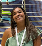 A young woman with long brown hair smiles wearing a mint-green off-the-shoulder ruffled blouse with a white lanyard around her neck while standing in front of a blue patterned staircase.