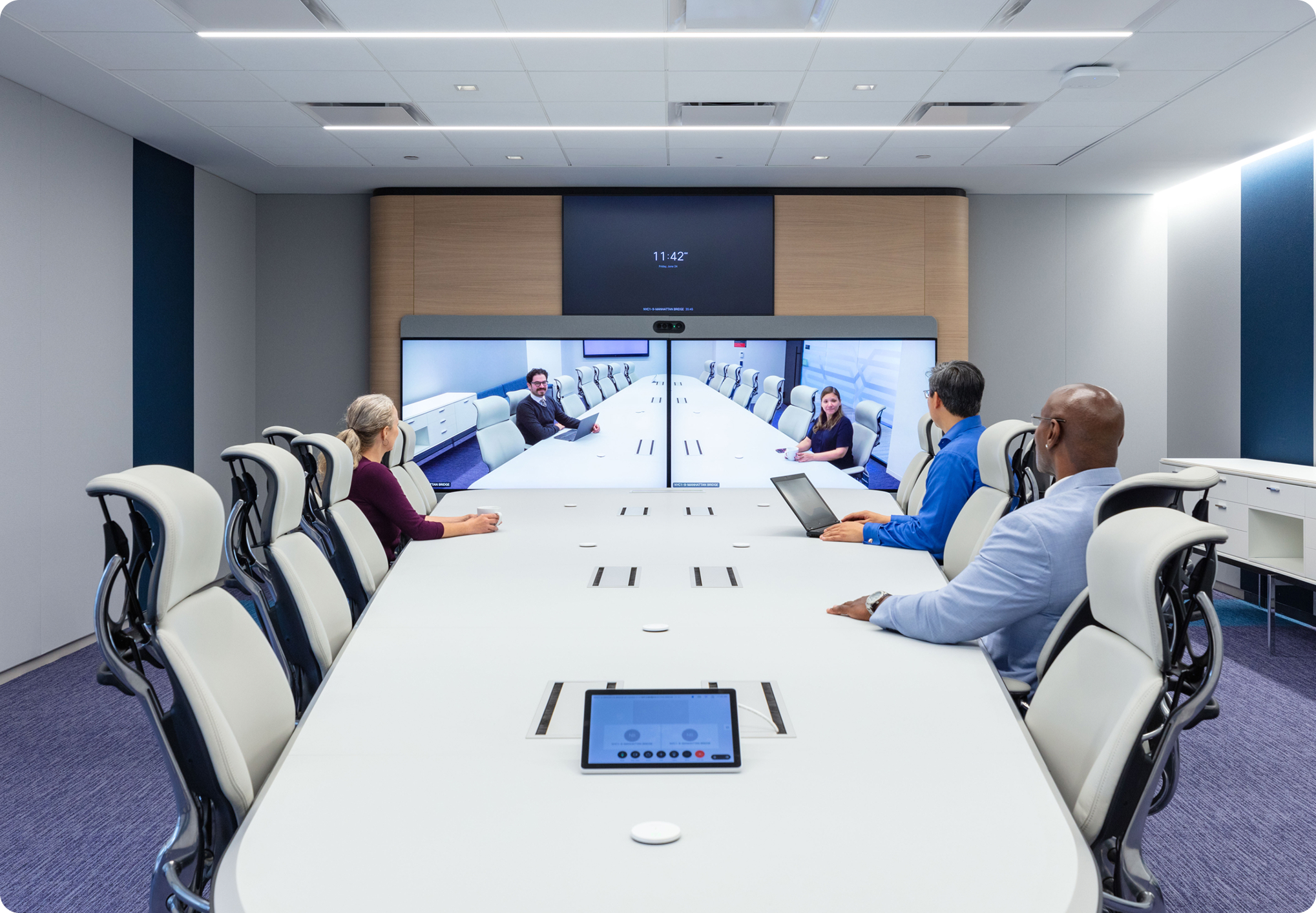 A modern conference room with people seated around the table looking at colleagues on large screens on a Webex call.