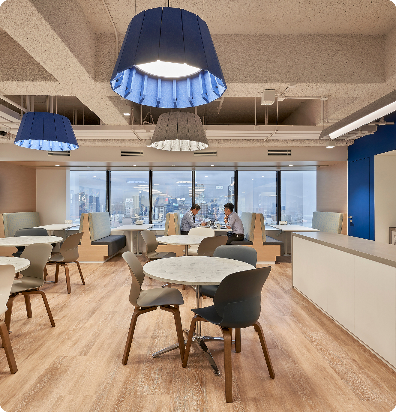 An open seating area with a number of tables and large, blue overhead lights. Two men sit at a table by the window working.