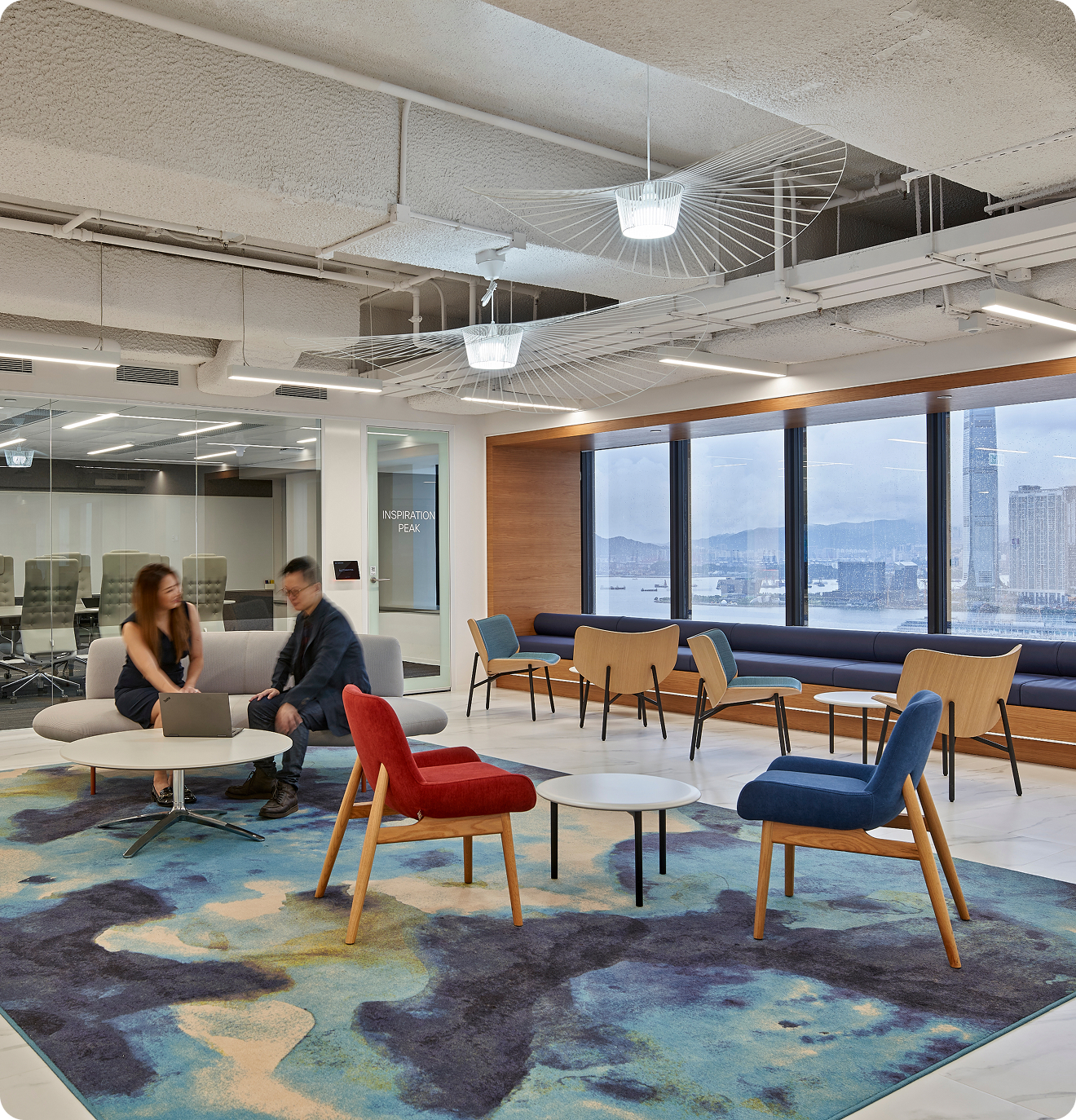 An open seating and work area with large windows looking out at a harbor. A woman and man sit on a couch looking at a laptop together.