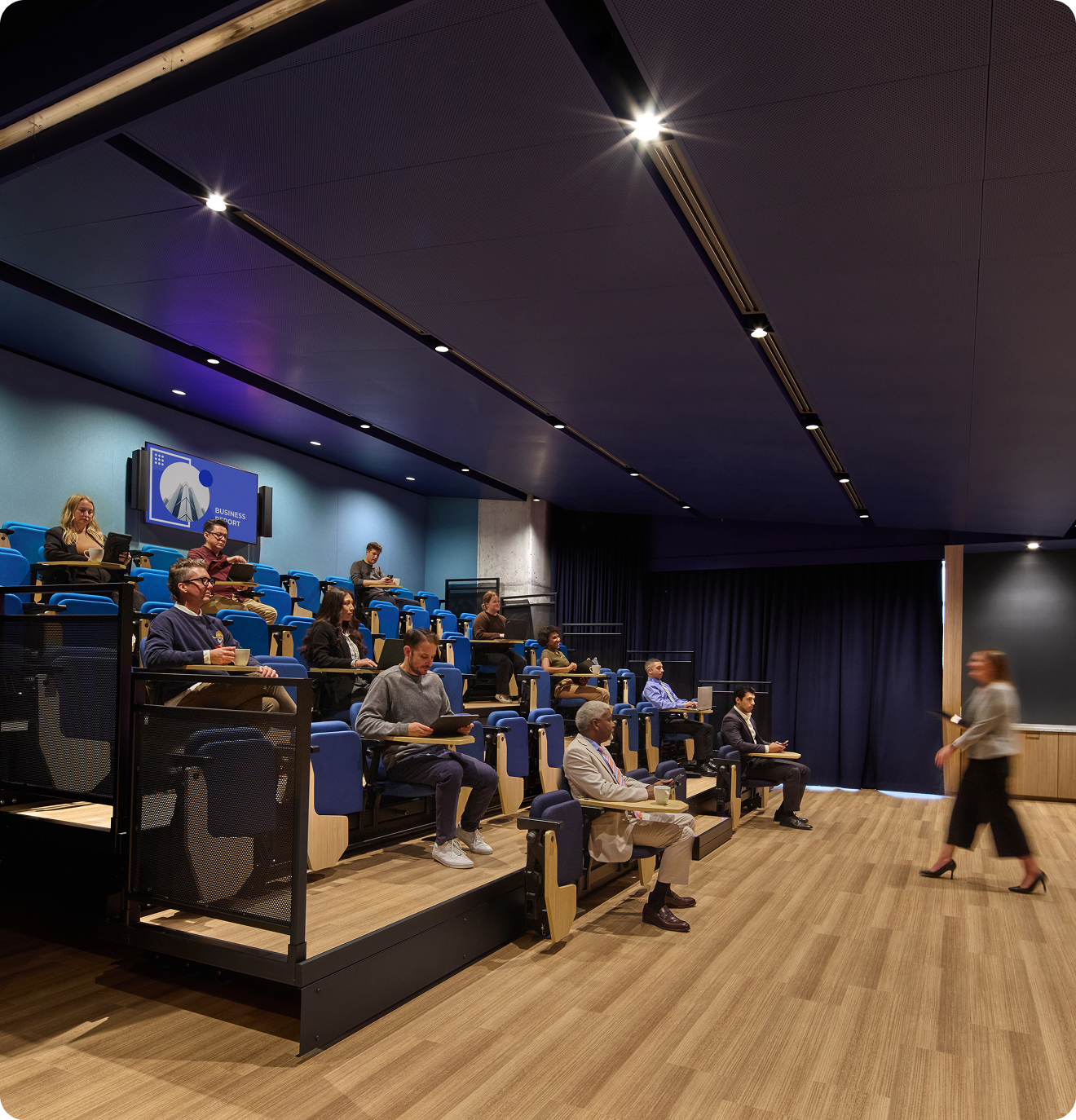 People sit in a theater space watching a woman giving a presentation.