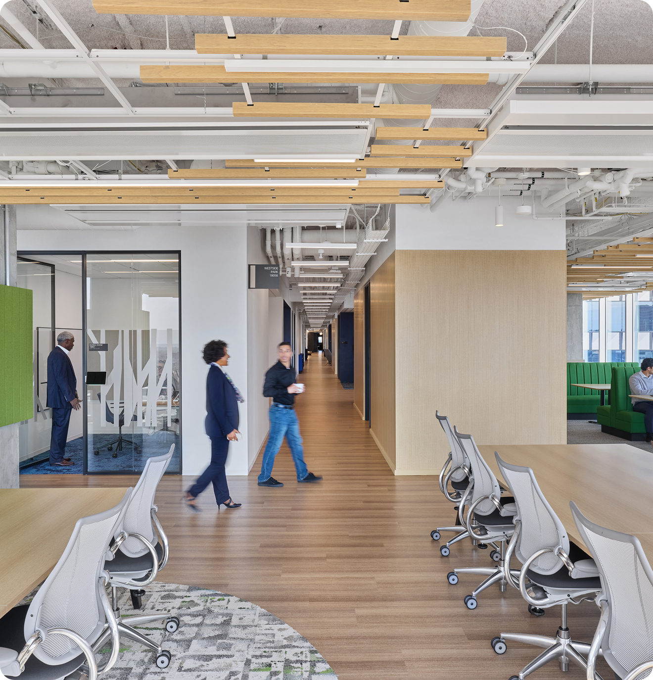 A light, airy workspace featuring long tables with chairs and conference rooms off to the sides with people walking through the space.