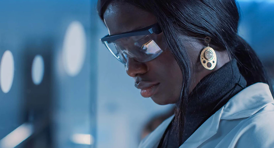 A woman in a medical environment wearing a lab coat and protective glasses.