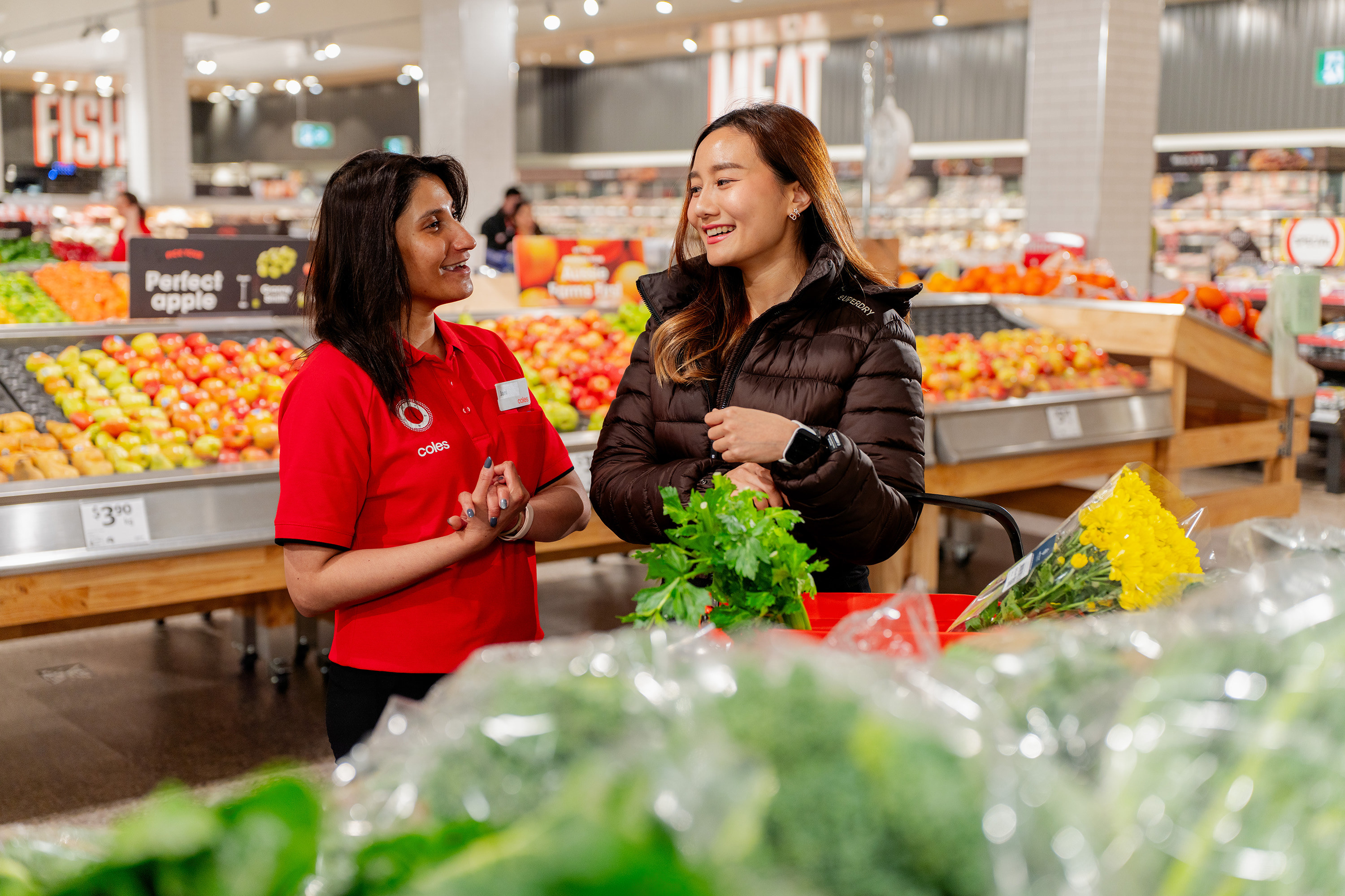A supermarket employee talks with a customer beside displays of fresh produce and flowers.