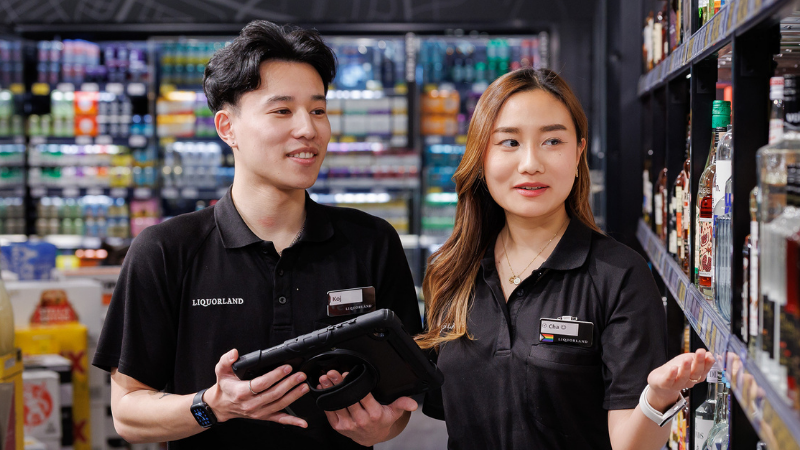 Two liquorland team members working together in store. A male team member to the left holds a tablet while the female team member to the right points to the shelves