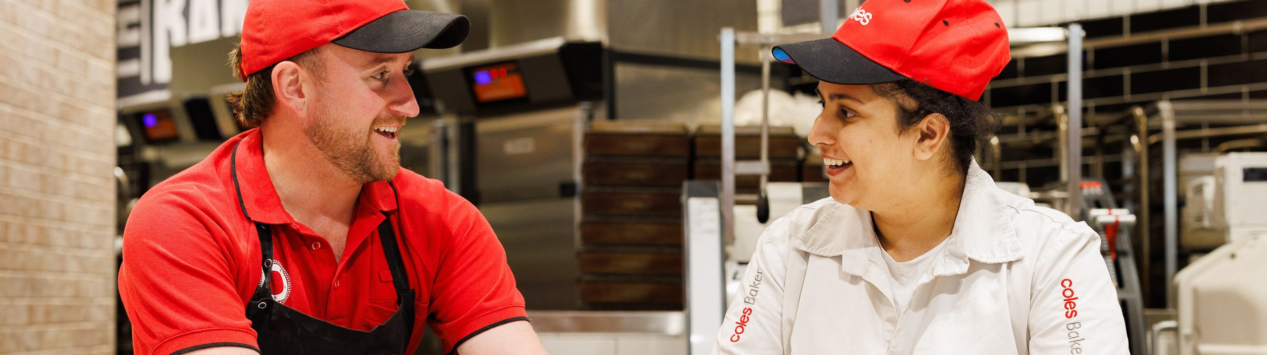 Two Coles bakers working together in the bakery. The male baker, smiling, wears a red polo, a black Coles-branded apron, and a red cap. The female baker, also smiling, wears a white Coles-branded uniform and a red cap.