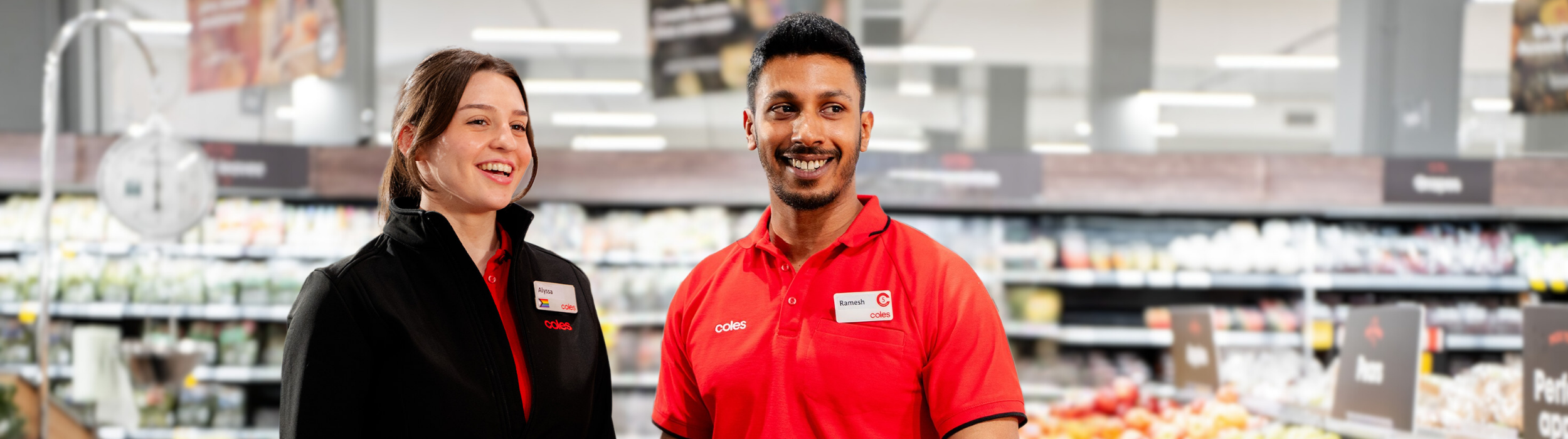 Two team members standing in fresh produce section of a Coles Supermarket. One team member wears a black branded Coles jacket while the other wears a Coles branded red polo.