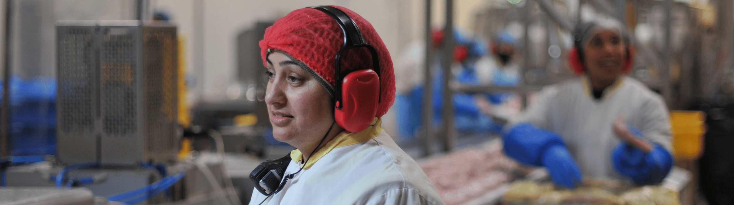 Workers at the Coles RROA facility wearing protective gear, including red hairnets, white coats, and red earmuffs, operating in a food processing environment with industrial equipment and conveyor belts in the background.