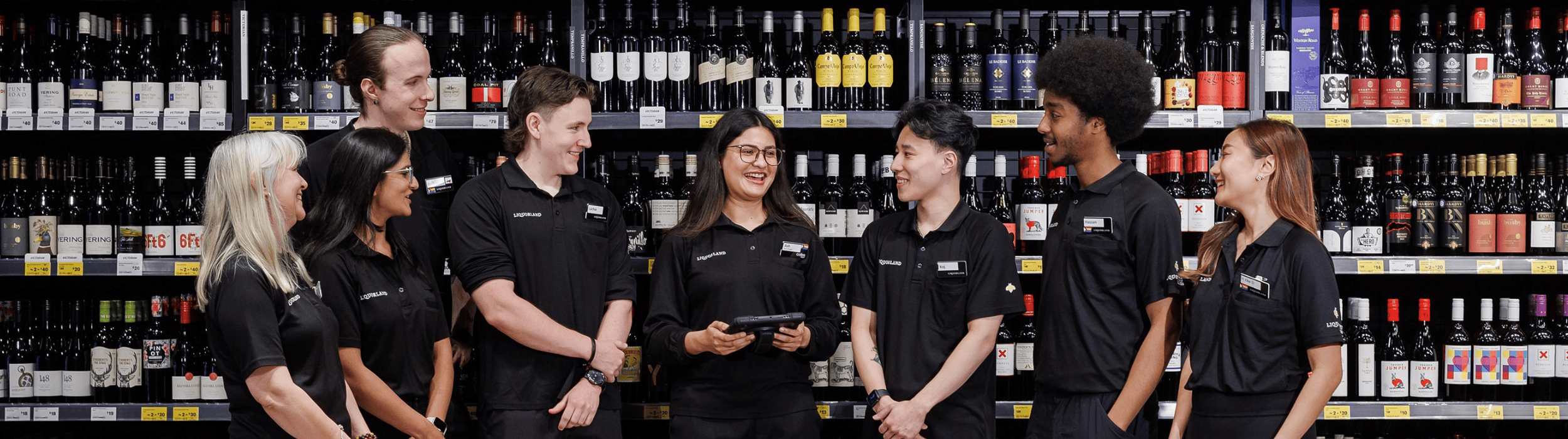 A group of various Liquorland team members standing instore in a team member huddle. The female team member in the center holds a tablet