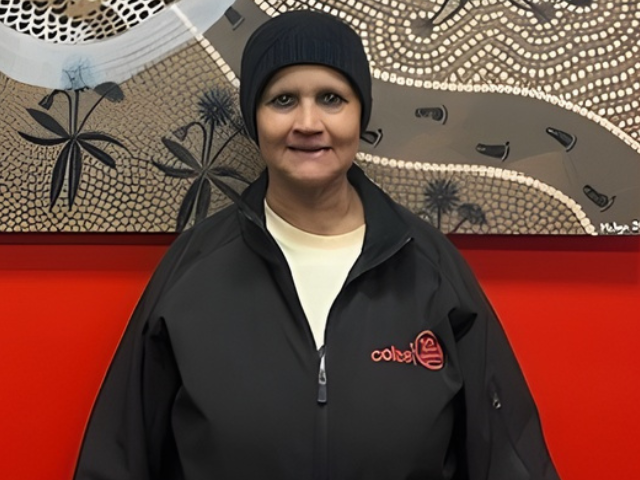 A female RROA team member in dark Coles jacket  and beanie stands in front of an Indigenous‑style artwork on a red wall, with an information plaque to the right.