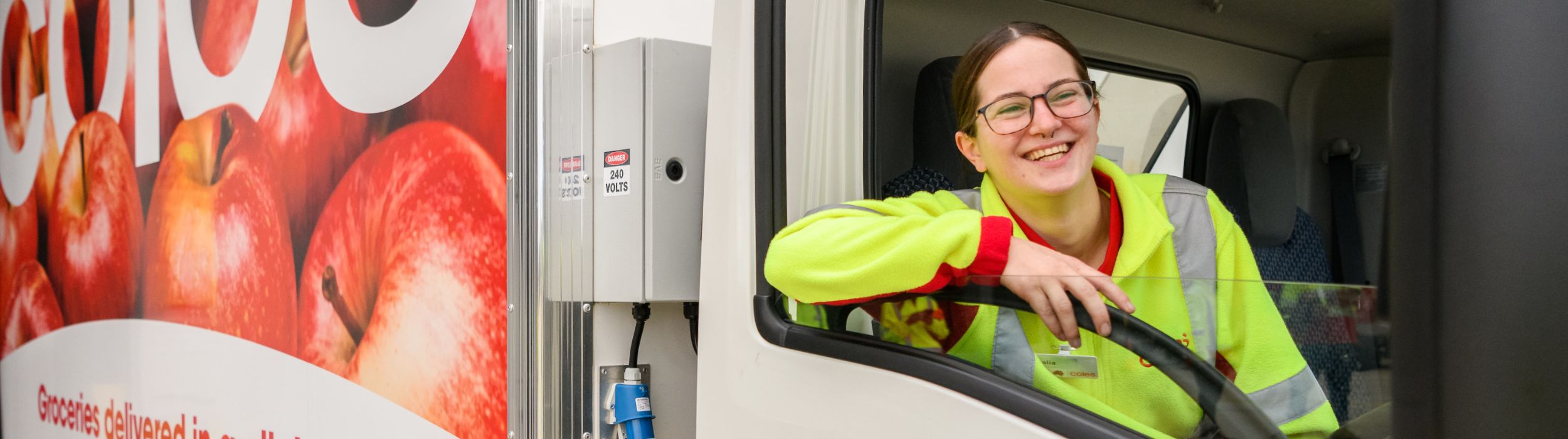 Female delivery driver in red polo and yellow high-visibility jacket  looking out the window while sat inside the Coles delivery van