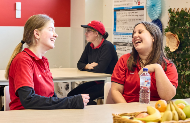 Two young store team members, one with dark blonde hair and another with brunette hair, sharing a light-hearted moment and engaging in conversation while seated together in the lunchroom.