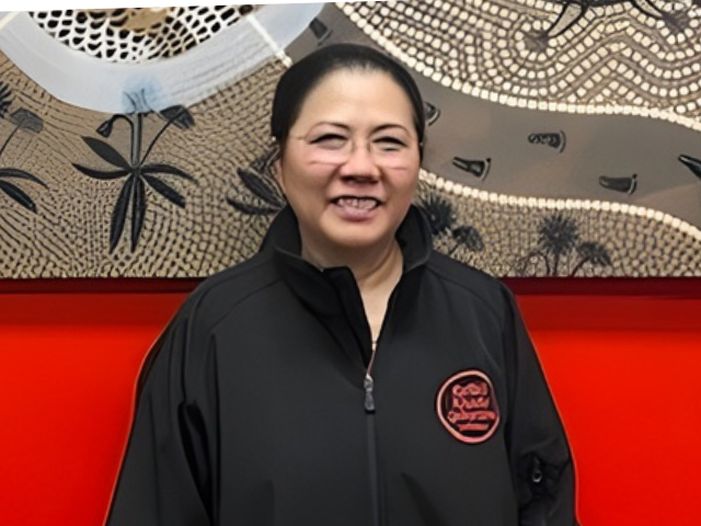 A female RROA team member in dark Coles jacket stands in front of an Indigenous‑style artwork on a red wall, with an information plaque to the right.
