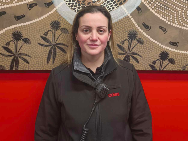 A woman in a dark Coles jacket stands in front of a large Indigenous‑style artwork on a red wall.