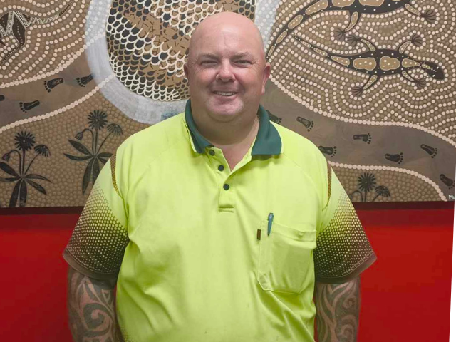 A male RROA team member wearing a high‑visibility yellow shirt stands in front of an Indigenous‑style artwork on a red wall.