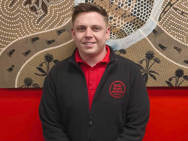 A man wearing a red shirt and dark jacket stands in front of a large Indigenous‑style artwork on a red wall, with an information plaque to the right.