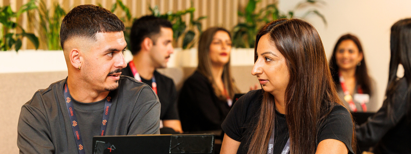 Two colleagues sitting side by side in a modern office lounge area, engaged in conversation with laptops in front of them. Other team members are visible in the background, creating a collaborative and open work environment with greenery and wooden accents.