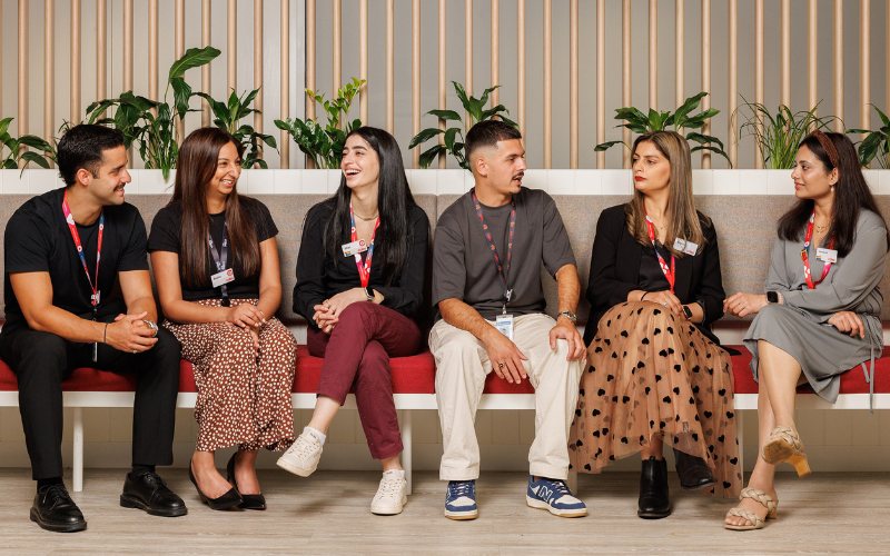 A diverse group of team members sitting together on a modern red bench in a bright, welcoming office space with green plants in the background. They are wearing workplace lanyards and engaging in conversation, reflecting collaboration and inclusion.