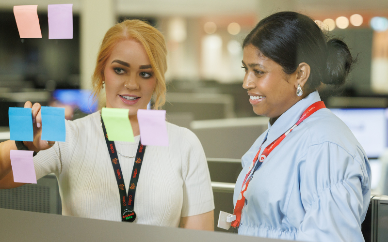 Two team members collaborating in an office space, reviewing colourful sticky notes on a glass wall. The scene reflects brainstorming and teamwork in an open-plan environment.