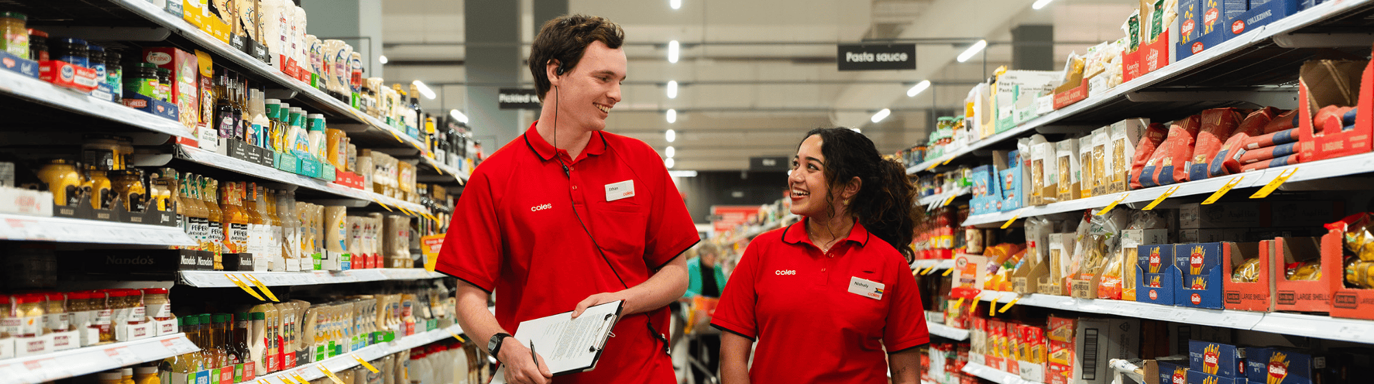 A young team member walking down a Coles supermarket aisle with a senior team member. Both are wearing signature Coles red polos