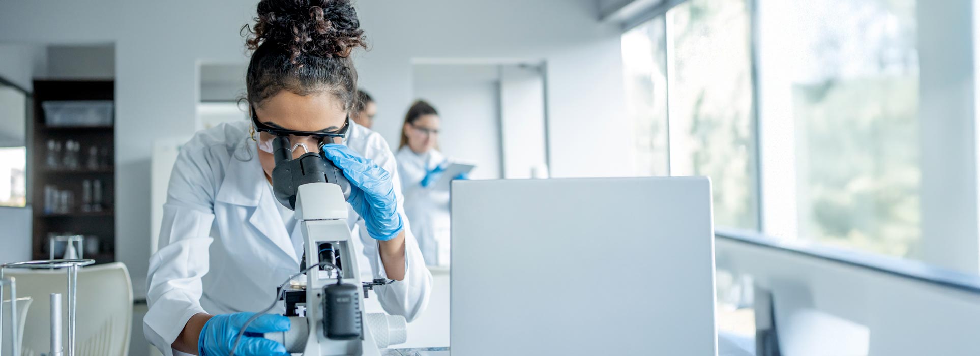 Female employee in lab coat and protective equipment looking into microscope