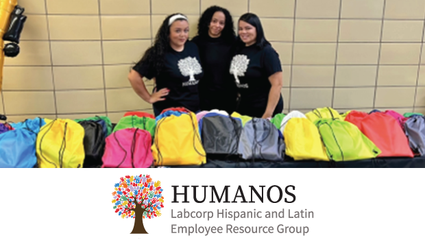 Three members of the HUMANOS ERG stand behind a table filled with colorful drawstring backpacks during a back-to-school drive. Collected over 100 backpacks and school supplies for Girls Prep Bronx and Martin Luther King Jr. School in Jersey City.