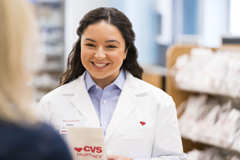 A smiling pharmacist wearing a white lab coat and nametag stands at a counter in a CVS Pharmacy, holding a paper bag with the CVS Pharmacy logo. Shelves of products are visible in the background. Another person, seen from behind, interacts with the pharmacist.