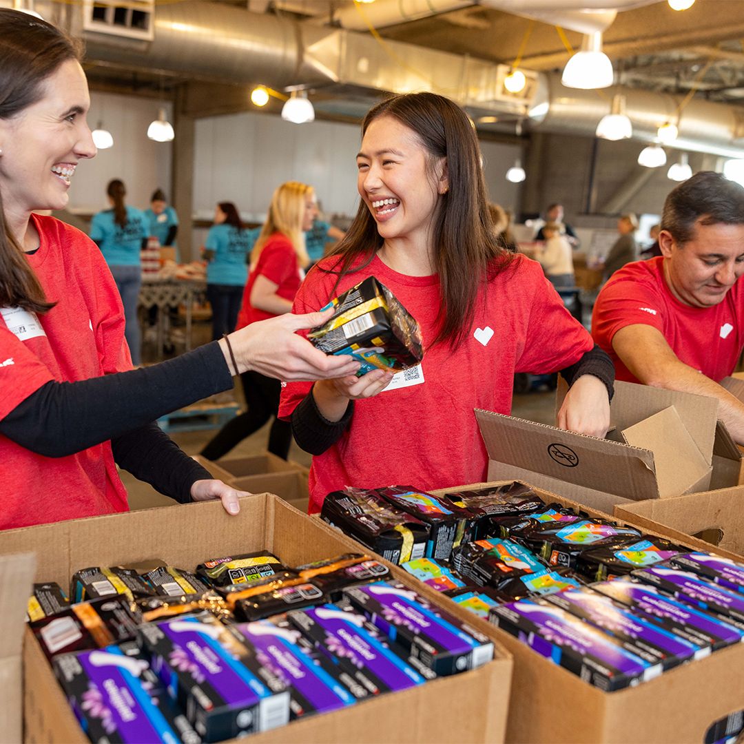 Volunteers wearing red shirts smile and work together while packing boxes with various supplies in a bustling warehouse setting. Other volunteers can be seen working in the background, wearing blue shirts. The atmosphere appears collaborative and cheerful.