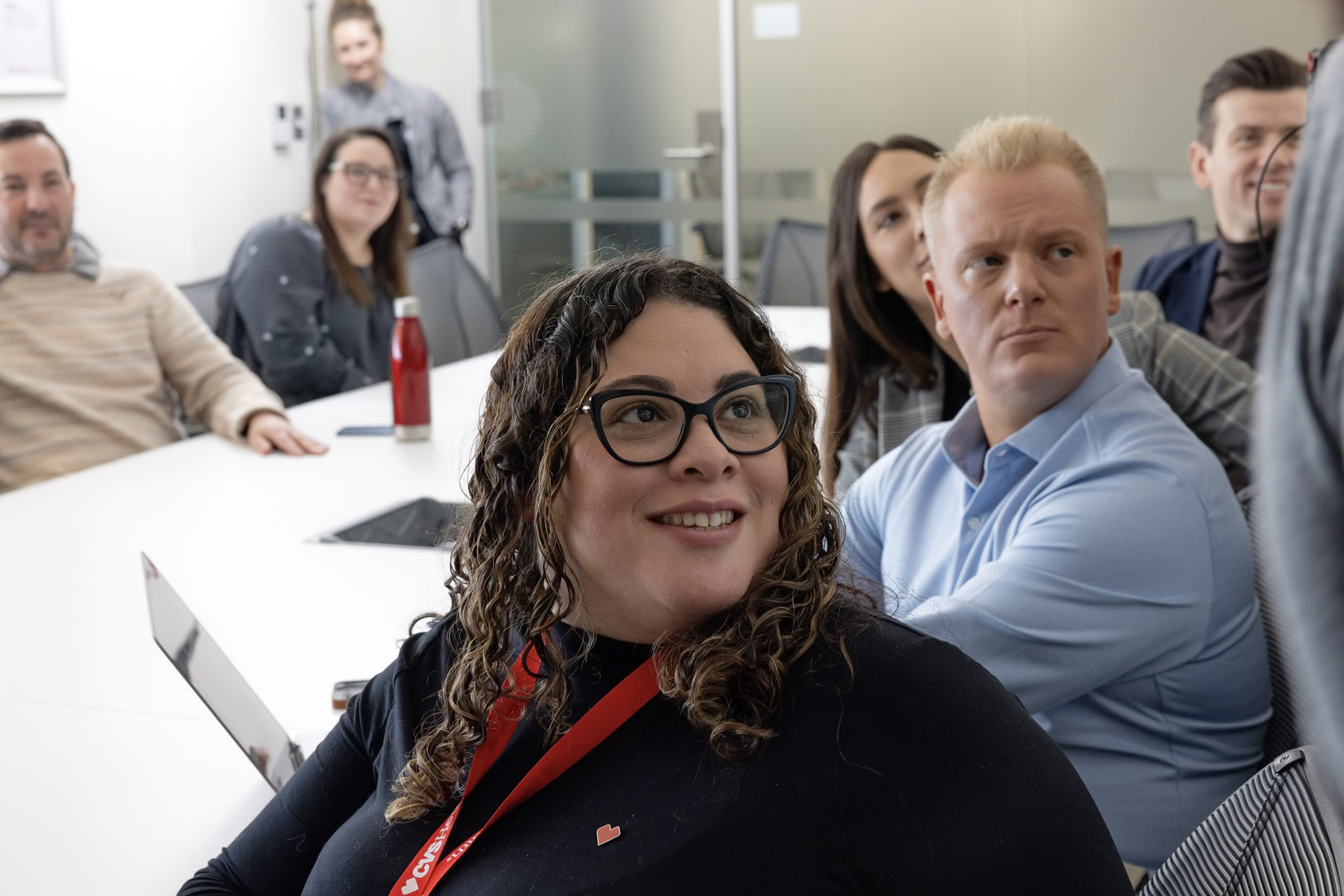 A group of office workers is gathered in a conference room. A woman with curly hair and glasses, sitting at the table with a laptop, smiles and looks towards someone off-camera. Others in the room are attentively looking in the same direction.