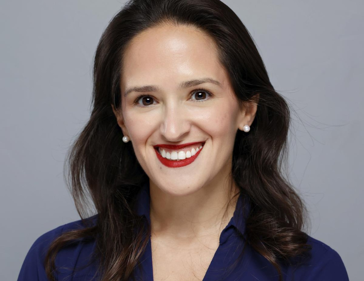 Alexandra wearing a deep blue collared shirt, with shoulder‑length dark hair styled with soft waves. The person is standing against a plain light gray background.