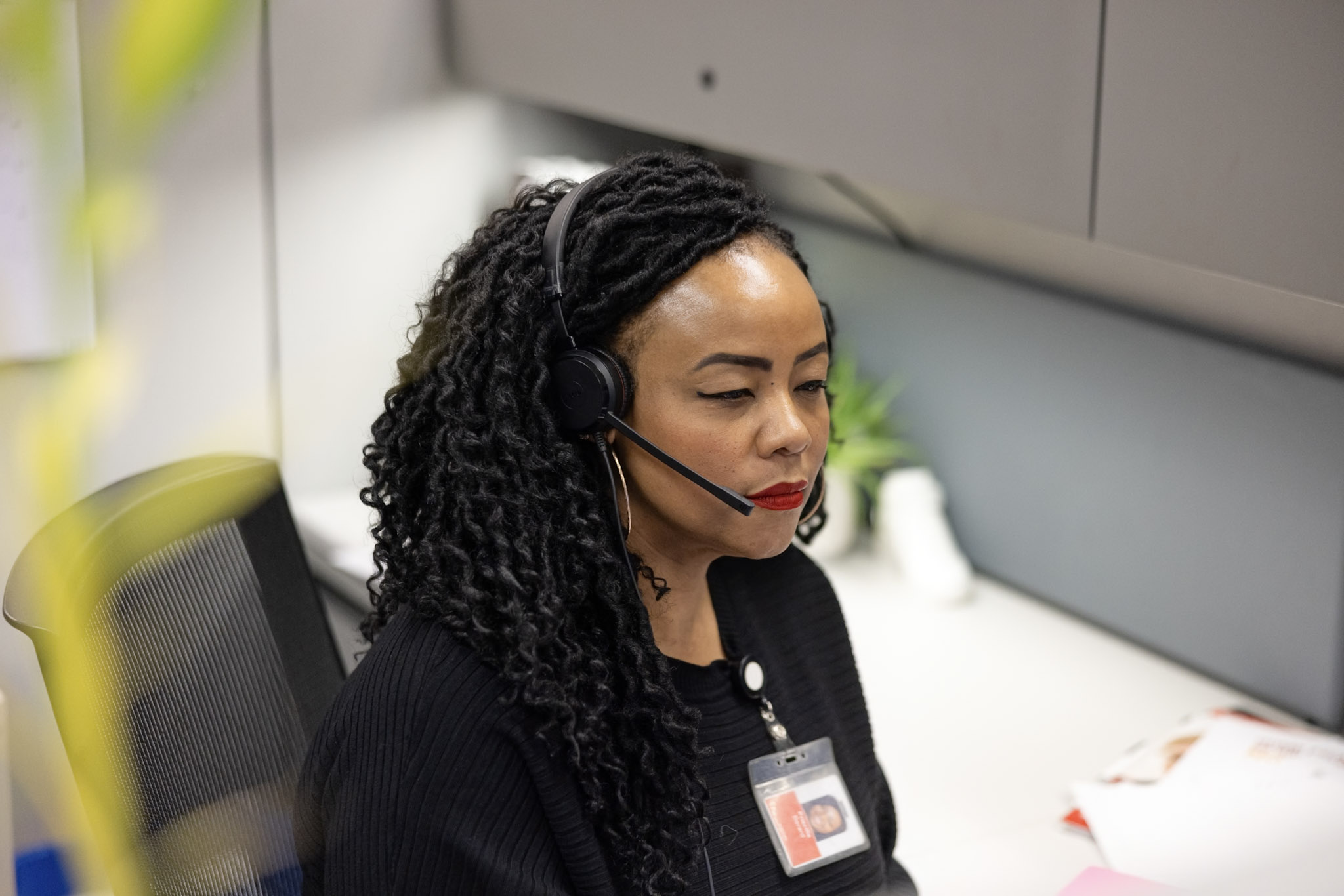 A person with long curly hair, wearing a headset and black sweater, sits at a desk with a name badge around their neck. They appear focused on their work. The background includes office furniture and a plant.