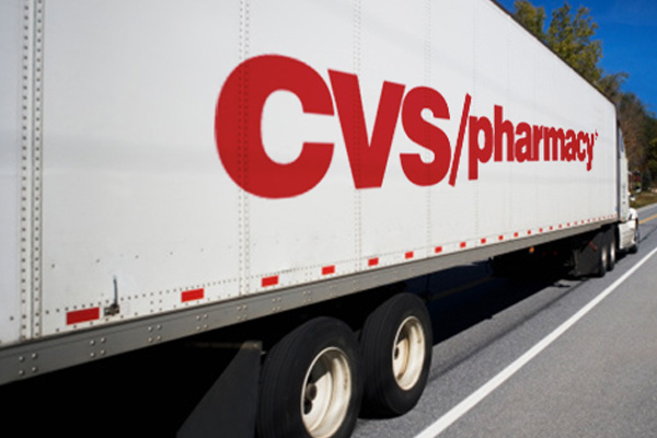 A large white truck with the red "CVS/pharmacy" logo on its side is driving on a road. The truck is moving forward, with trees and a clear sky visible in the background.