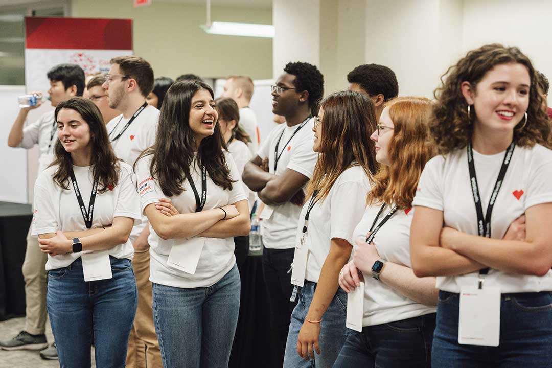 A diverse group of young adults wearing white T-shirts and lanyards stand together in a room. Some are chatting and smiling while others look ahead. They appear to be participating in an event or conference. In the background, a person is drinking from a water bottle.