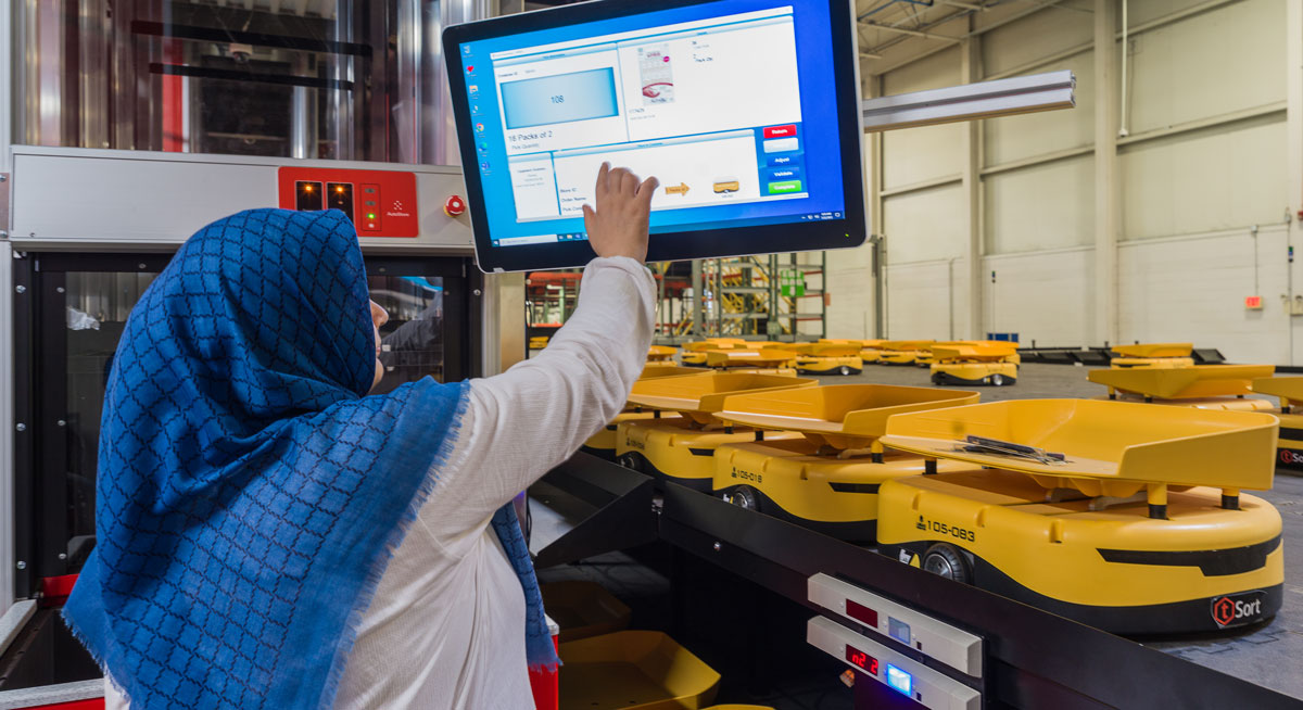 A person wearing a blue patterned headscarf and a white shirt operates a touch-screen monitor in a warehouse setting. Numerous yellow automated guided vehicles (AGVs) are seen on a conveyor system in the background.