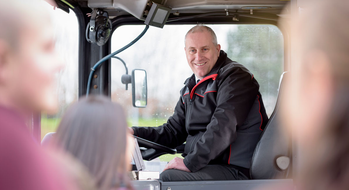 A smiling bus driver wearing a black and red jacket sits at the driver's seat, looking towards the camera. The blurred heads of passengers, including children, are visible in the foreground. The bus interior and driver's dashboard are partially shown.