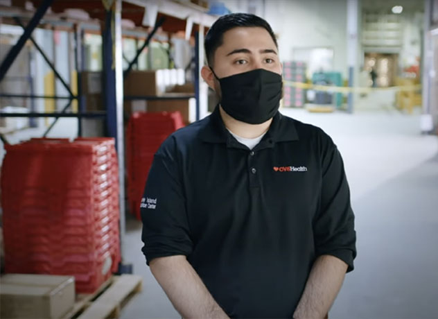 A person wearing a black mask and a black shirt with a "CVS Health" logo stands in a warehouse. Shelves with red bins are visible behind them, alongside various equipment and supplies. The environment appears organized and functional.