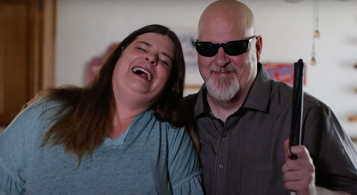 A woman with long brown hair and a blue shirt laughs while leaning on a man with a bald head, white beard, and sunglasses. The man, holding a white cane, smiles while standing next to her. They appear to be in a casual indoor setting.